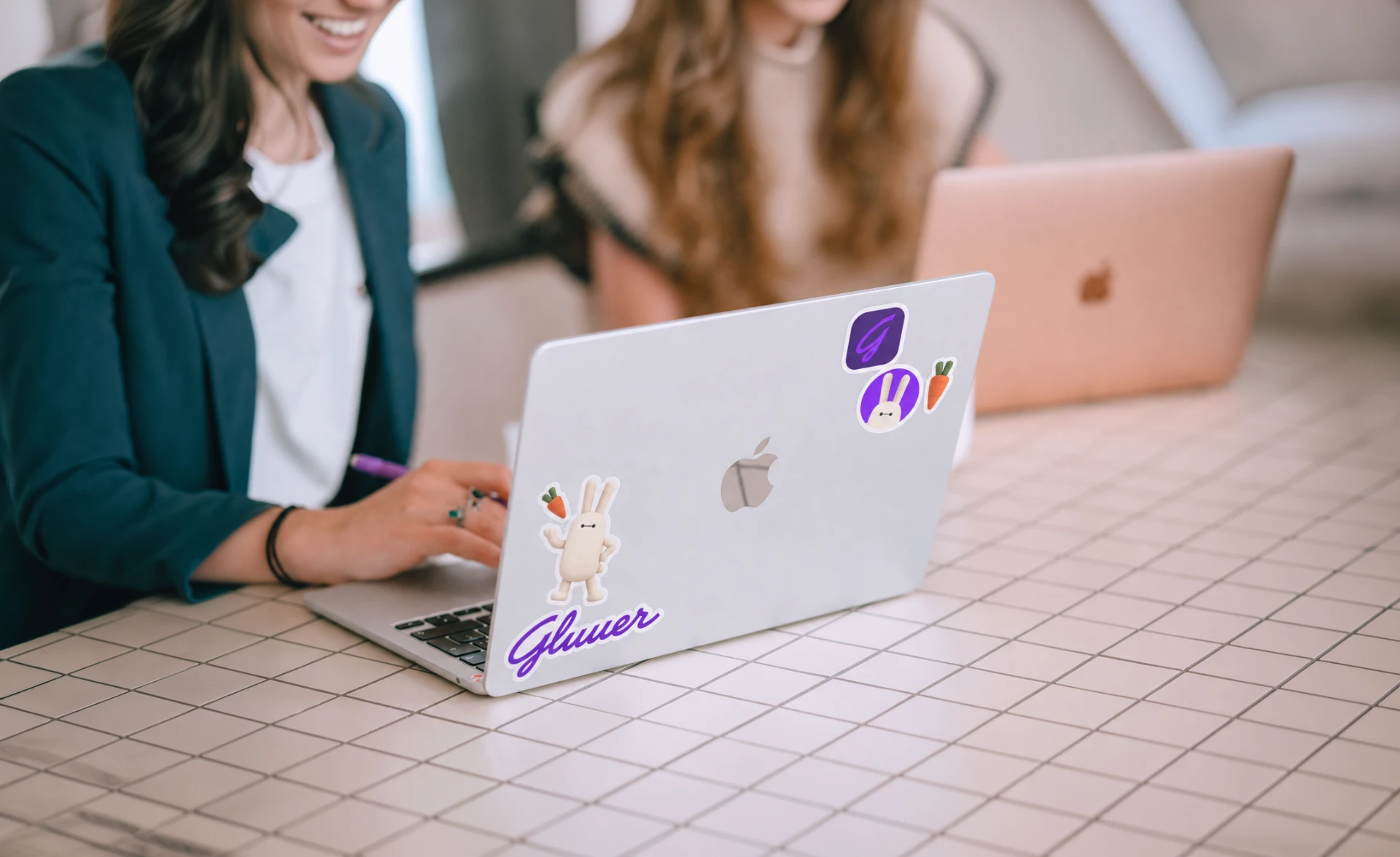 A silver laptop on a grid-patterned table decorated with Gluuer stickers, including the purple Gluuer wordmark and the cream rabbit mascot holding a carrot, while a smiling woman works at the keyboard in a bright room